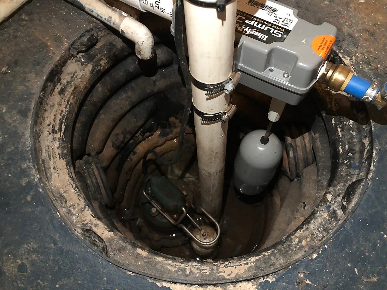 A sump pump sitting in its basin in a basement floor, with the discharge pipe running up and a backup pump mounted above